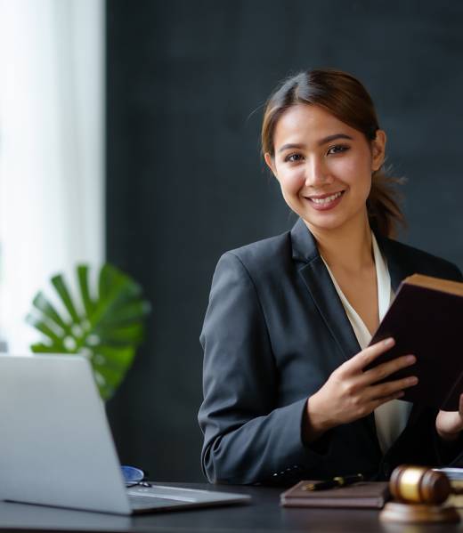 Beautiful Asian woman lawyer sitting at a table smiling happy with a laptop computer with law books hammer and scales hammer and scale.